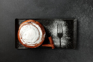 Round, delicious bun with cinnamon and powdered sugar on a black plate on a gray background