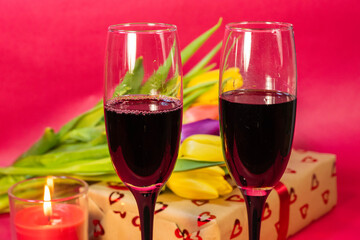 a romantic celebration featuring two champagne flutes filled with red wine, a heart-patterned gift box, fresh tulips, and a lit candle on a red background.