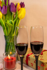 a romantic setting featuring two glasses of red wine, a heart-patterned gift box, and a vase of colorful tulips illuminated by a lit red candle.