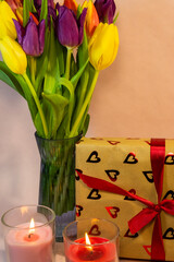 a romantic display of colorful tulips in a glass vase, a heart-patterned gift box, and two lit candles set against a beige background.