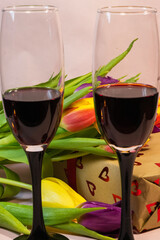 two glasses filled with red wine standing in front of a heart-patterned gift box and a colorful bouquet of fresh tulips.