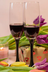 two glasses of red wine, a lit red candle, and a heart-patterned gift box nestled among fresh colorful tulips.