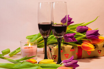 two glasses of red wine, a lit red candle, and a heart-patterned gift box nestled among fresh colorful tulips.
