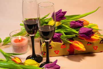 two glasses of red wine, a lit red candle, and a heart-patterned gift box nestled among fresh colorful tulips.