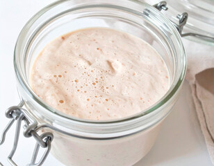 Close-up view of a sourdough starter in a glass jar, showing an active, aerated surface with small bubbles. Natural fermentation mixture prepared for bread making, photographed in neutral light.