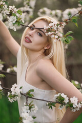 A beautiful girl with long blond hair in a blooming garden, a portrait among branches of apricot and cherry blossoms. Spring is the blossoming of femininity, beauty, and peace.