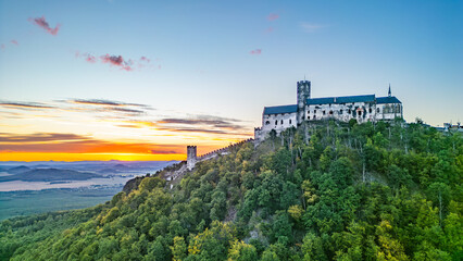 Stunning aerial view of Bezdez Castle perched atop a hill, surrounded by lush greenery, showcasing its medieval towers against a vibrant sunset sky during a summer evening.
