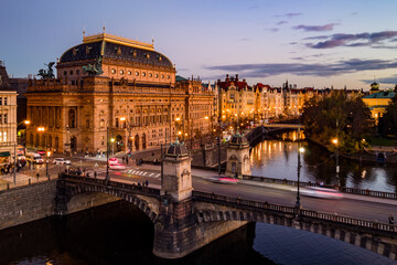 Aerial view of the National Theatre in Prague at evening. The theatre is illuminated against a darkening sky, with the Vltava River and embankment visible. Traffic moves across the bridge.
