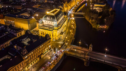The National Theatre is lit up in the evening sky above the Vltava River in Prague. Streets are busy with cars and people. Bridges connect the banks on this vibrant night.