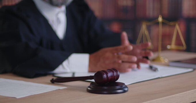 Judge gavel lying on desk with law and justice balance scales, legal paper documents in courtroom. Hands of lawyer clasped at desk, magistrate in robe delivering decision and verdict during trial