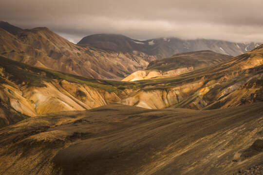 View of rugged, colorful mountains and valleys under a dramatic sky, where golden hues meet earthy browns, creating a painterly landscape, Landmannalaugar, Central Highland, Iceland.