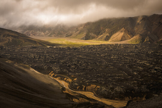 View of rugged black lava fields stretch towards distant mountains under a moody sky, contrasting with a patch of vibrant green, Landmannalaugar, Iceland.