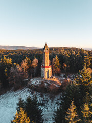 Stepanka lookout tower stands tall in the Jizera Mountains during winter. Sunlight shines on the tower surrounded by snow-covered trees and mountains in the background.