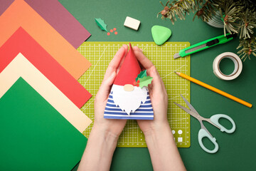 Hands holding a finished gnome craft made of colored paper. Flat lay of handmade holiday decorations, creative hobby, and master class concept.