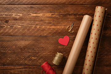 Top view flat lay of gift wrapping supplies on wooden table with kraft paper rolls, scissors, twine and red heart tag. Valentine copy space.