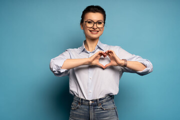 Smiling middle aged woman making heart shape with hands on blue background.