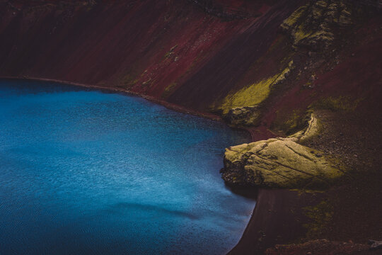 View of the serene blue water nestled against the stark contrast of the red and black volcanic landscape, with patches of green moss, Landmannalaugar, The Central Highland, Iceland.