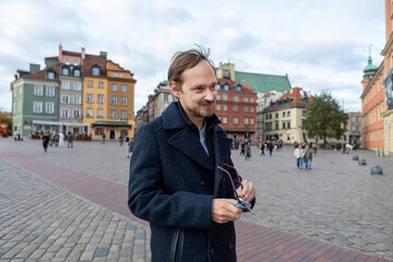 Young man smiles while holding glasses in a city square in Warsaw during the day