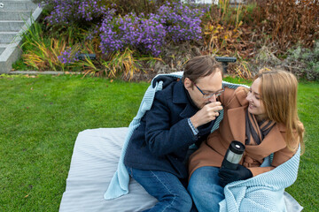 Couple enjoys warm drinks while sitting on a blanket in a garden setting during autumn season