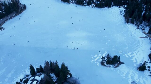Aerial drone footage of a frozen lake in Champex, Switzerland, during winter. People ice skating surrounded by snowy alpine scenery. Shot in 4K using a DJI Mini 2.