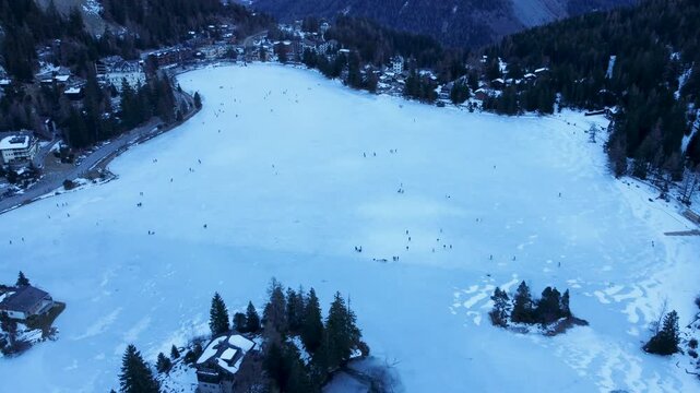 Aerial drone footage of a frozen lake in Champex, Switzerland, during winter. People ice skating surrounded by snowy alpine scenery. Shot in 4K using a DJI Mini 2.