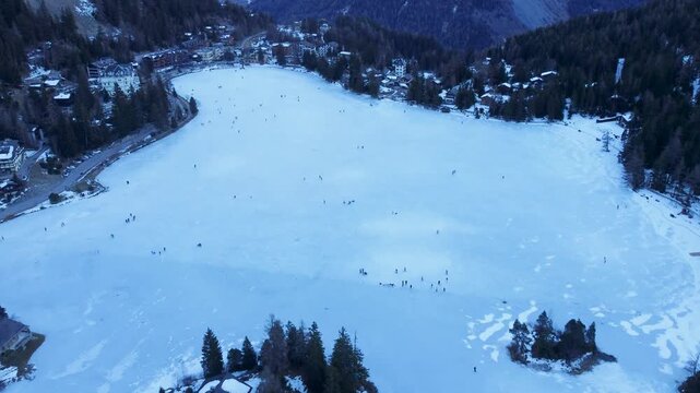 Aerial drone footage of a frozen lake in Champex, Switzerland, during winter. People ice skating surrounded by snowy alpine scenery. Shot in 4K using a DJI Mini 2.