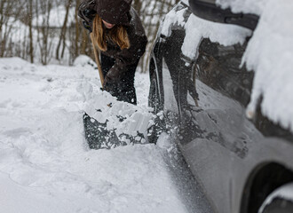 Woman with a snow shovel clears snow from a snow-covered car so she can drive out of the parking lot in front of her house