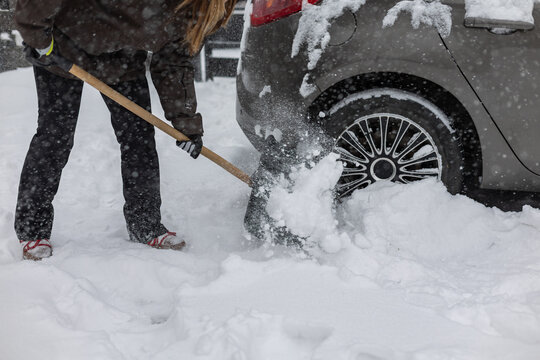 Woman with a snow shovel clears snow from a snow-covered car so she can drive out of the parking lot in front of her house
