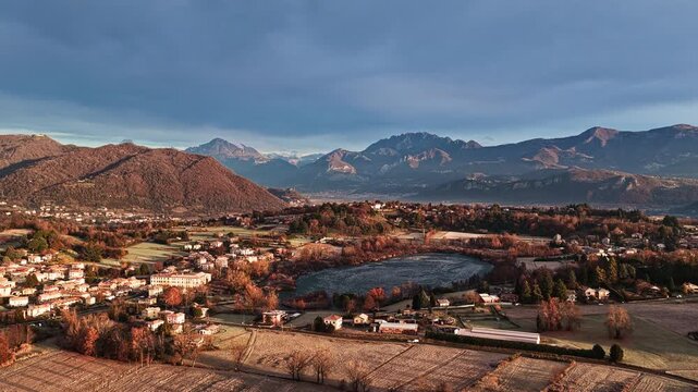 Aerial drone shot moving forward toward a frozen lake, revealing winter countryside, fields and mountains in northern Italy during a calm sunrise.