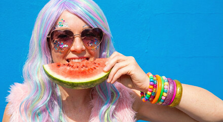 Eccentric woman with colorful hair and heart sunglasses eating watermelon slice against blue background
