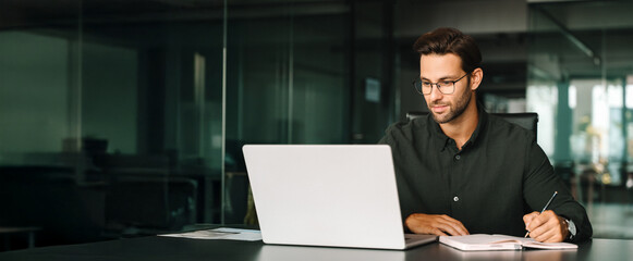 Young adult European man looking at laptop pc screen. Motivated latin student writing notes during...