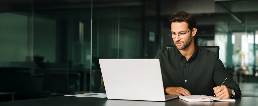 Young adult European man looking at laptop pc screen. Motivated latin student writing notes during lesson, watching webinar, learning online, sitting at work desk in office. Web banner, copy space