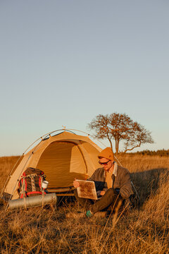 Traveling male freelancer typing on laptop while sitting near camping tent during remote work in nature  