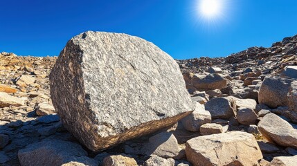 A large rough unworked granite boulder rests on a rocky mountainside under a clear blue sky with bright sunlight