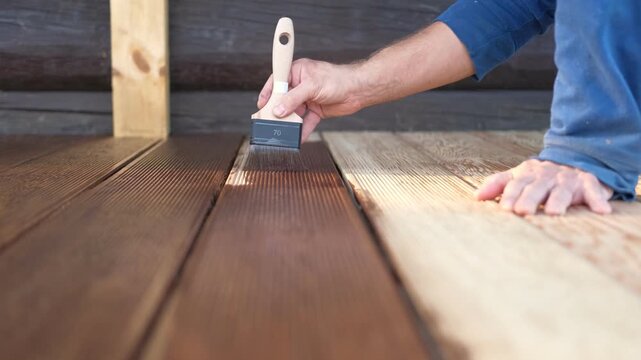 Brown oil for protection of wood by staining. Painting a larch for protecting and pests. Woodworker applying finish to terrace deck board. Repairman varnishing with brush wooden plank floor surface.