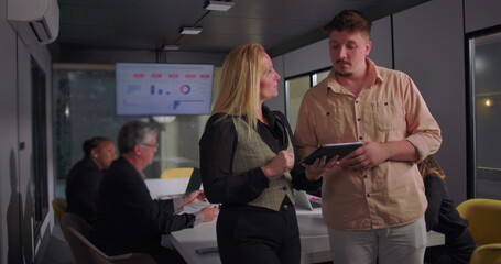 Young man and blonde businesswoman collaborating over digital tablet in office, team working in background with presentation data on monitor