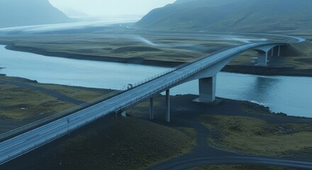 Modern concrete bridge spans a glacial river surrounded by misty mountains and green fields.