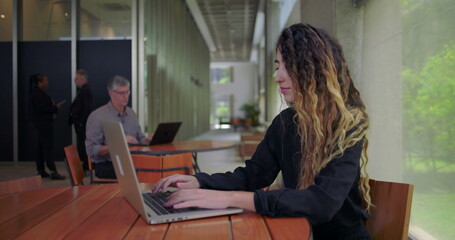 Woman typing on laptop at table in open office space while man and two others work and talk in background modern workplace environment
