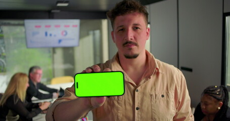 Young man presenting horizontal smartphone with green screen toward camera, serious expression in office setting with team working at conference table
