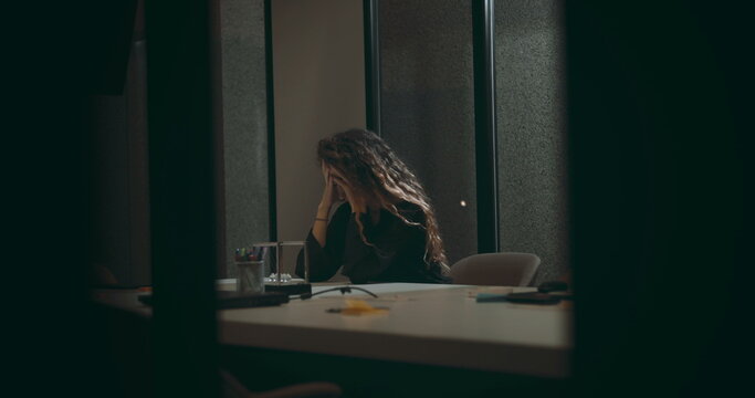 Young businesswoman sitting alone at office table holding head in frustration, overwhelmed by workplace stress and pressure after intense corporate meeting