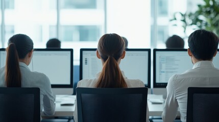 Three office workers sit at desks, working on computers in a modern, bright workspace.