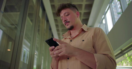 Young man using smartphone in office hallway, looking at screen with calm and focused expression, casual and modern workspace environment