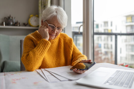 Senior woman holding mobile phone while reading paper documents at home
