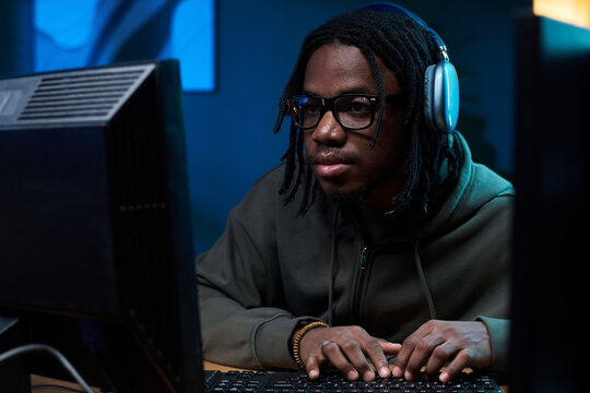 Young Black man wearing glasses and headphones sitting at desk using desktop computer, focusing intently on screen while typing on keyboard in workspace - Powered by Adobe