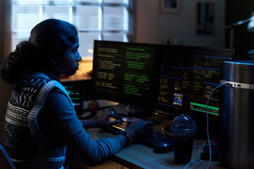 Young South Asian woman working at desk using multiple computer monitors with code displayed, typing on keyboard, focused on programming task in home office setting