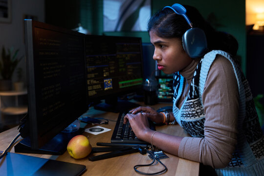 Young Asian woman wearing headphones working intently at computer with multiple monitors, typing on keyboard, surrounded by electronic devices and fruit on desk - Powered by Adobe