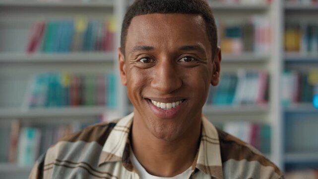 Happy African American man student smiling in library looking at camera friendly ethnic male guy learner teacher posing portrait books bookcase education studying university academy graduate knowledge