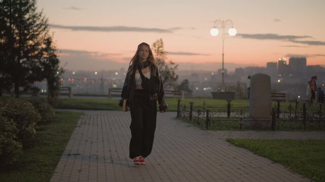caucasian woman walking at dusk along urban park pathway, sunset skyline and lamp glow, red sneakers and stylish coat, confident stride toward camera, cinematic evening atmosphere