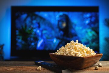 A wooden bowl of popcorn and remote control in the background the TV works. Evening cozy watching a movie or TV series at home