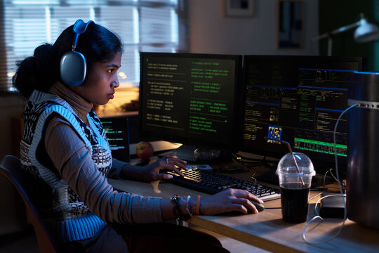 Young South Asian woman wearing headphones working on multiple computer monitors displaying code, sitting at desk with drink and apple, focusing intently on programming tasks
